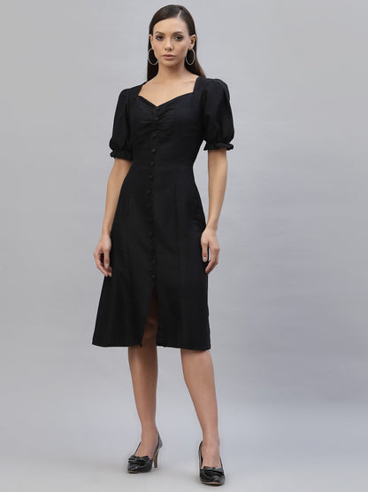 A woman wearing a black knee-length dress with short puffed sleeves and a V-neckline, standing against a grey background.