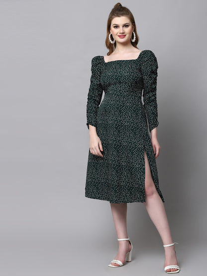 A woman standing against a grey background, wearing a green knee-length A-line dress with long sleeves and a square neckline.